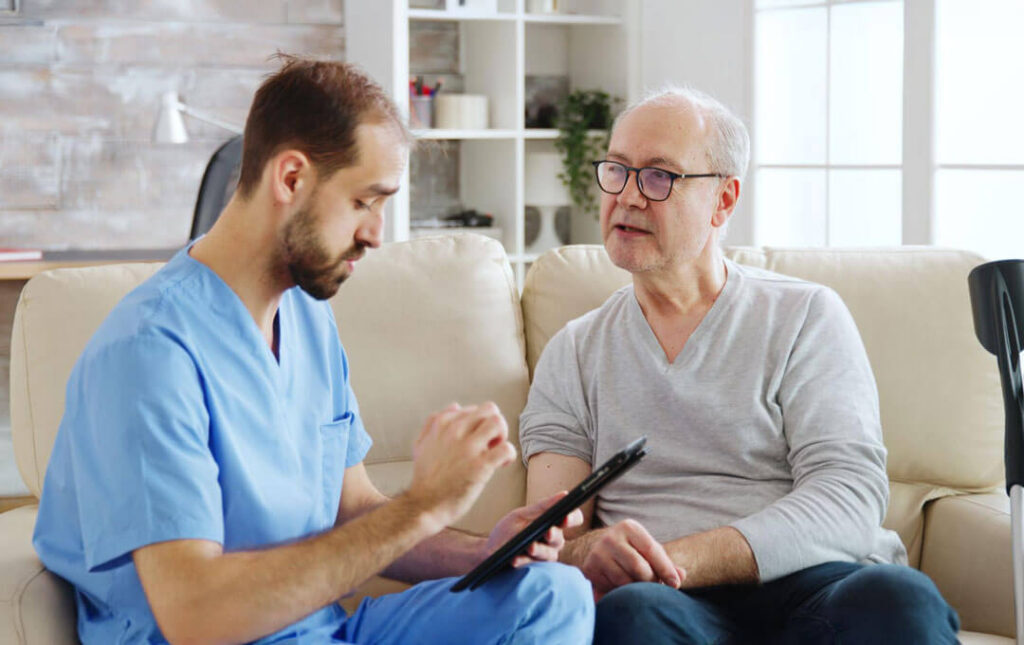A male healthcare provider in blue scrubs holds a tablet and talks with an older man seated beside him on a cream sofa in a bright, plant-filled living room.
