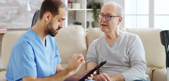 A male healthcare provider in blue scrubs holds a tablet and talks with an older man seated beside him on a cream sofa in a bright, plant-filled living room.