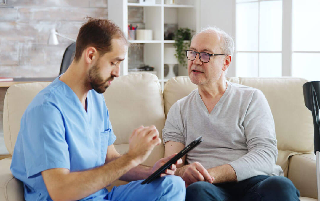 A male healthcare provider in blue scrubs holds a tablet and talks with an older man seated beside him on a cream sofa in a bright, plant-filled living room.