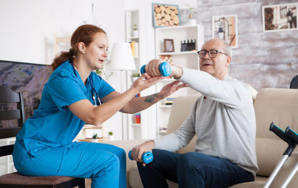 A nurse helps an older man do arm exercises with hand weights during a physical therapy session at home.