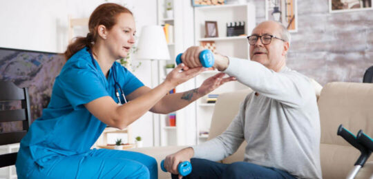 A nurse helps an older man do arm exercises with hand weights during a physical therapy session at home.