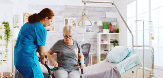 A nurse helps an elderly woman stand up from a bed using a walker in a care facility room.