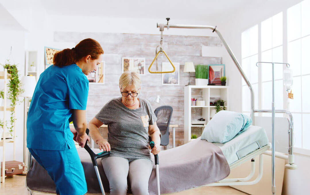 A nurse helps an elderly woman stand up from a bed using a walker in a care facility room.