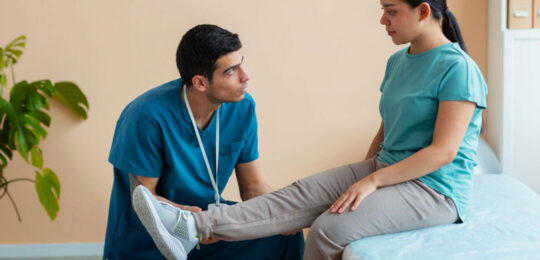 A male physical therapist in teal scrubs kneels on the floor and gently lifts the leg of a young woman seated on a treatment table, assessing her range of motion in a soft-toned rehabilitation room.