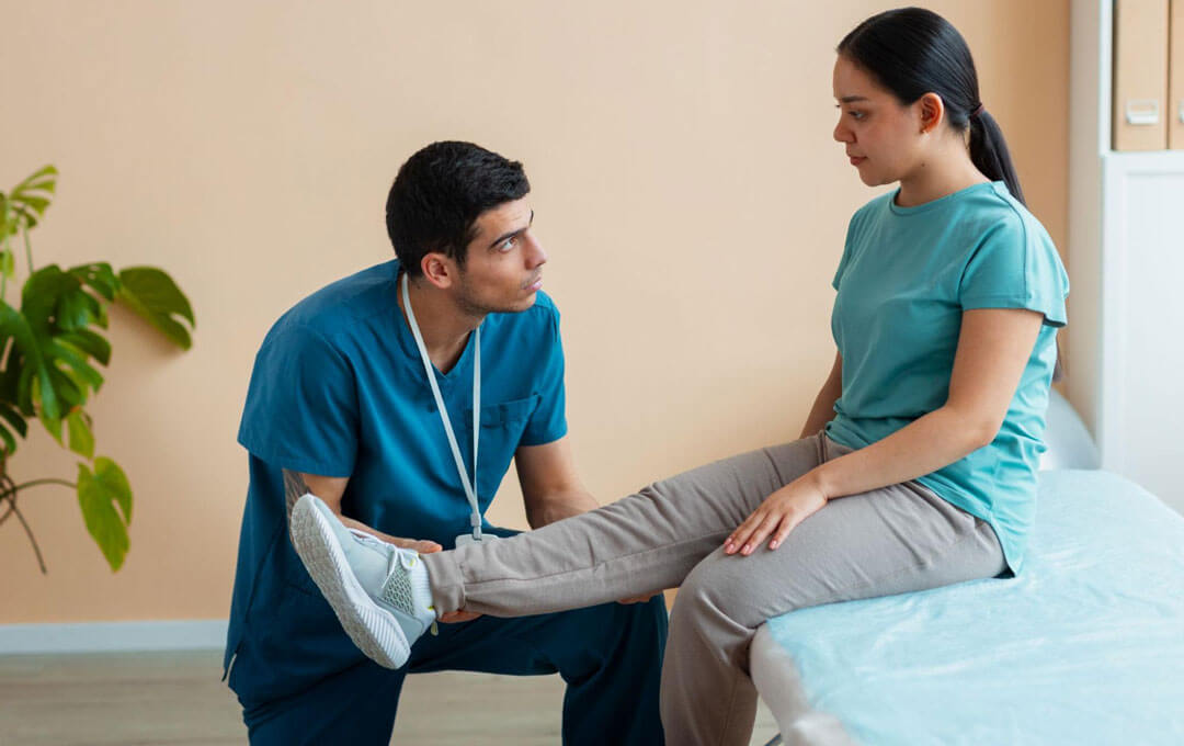 A male physical therapist in teal scrubs kneels on the floor and gently lifts the leg of a young woman seated on a treatment table, assessing her range of motion in a soft-toned rehabilitation room.