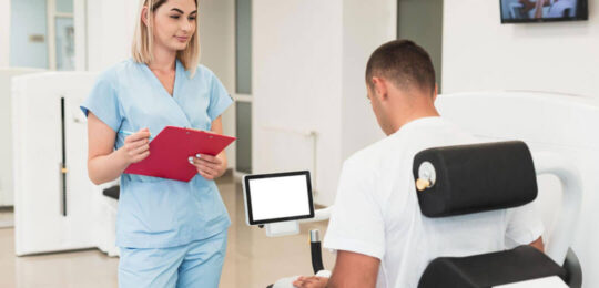 A female nurse in light blue scrubs holding a red clipboard stands and speaks with a male patient seated in a rehabilitation chair inside a bright, modern clinical facility.
