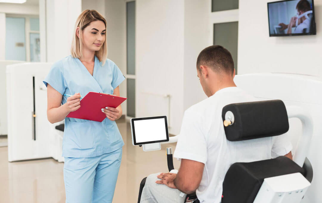 A female nurse in light blue scrubs holding a red clipboard stands and speaks with a male patient seated in a rehabilitation chair inside a bright, modern clinical facility.