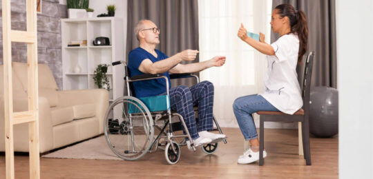 A rehabilitation therapist guides a senior man in a wheelchair through an upper-body mobility exercise.