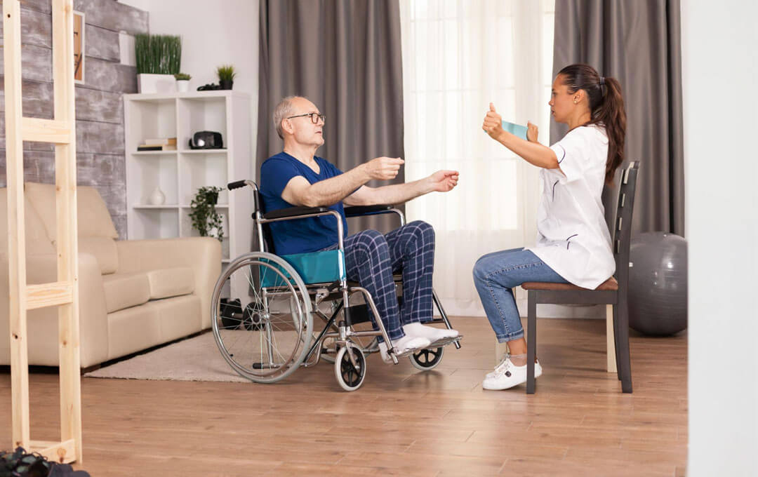 A rehabilitation therapist guides a senior man in a wheelchair through an upper-body mobility exercise.